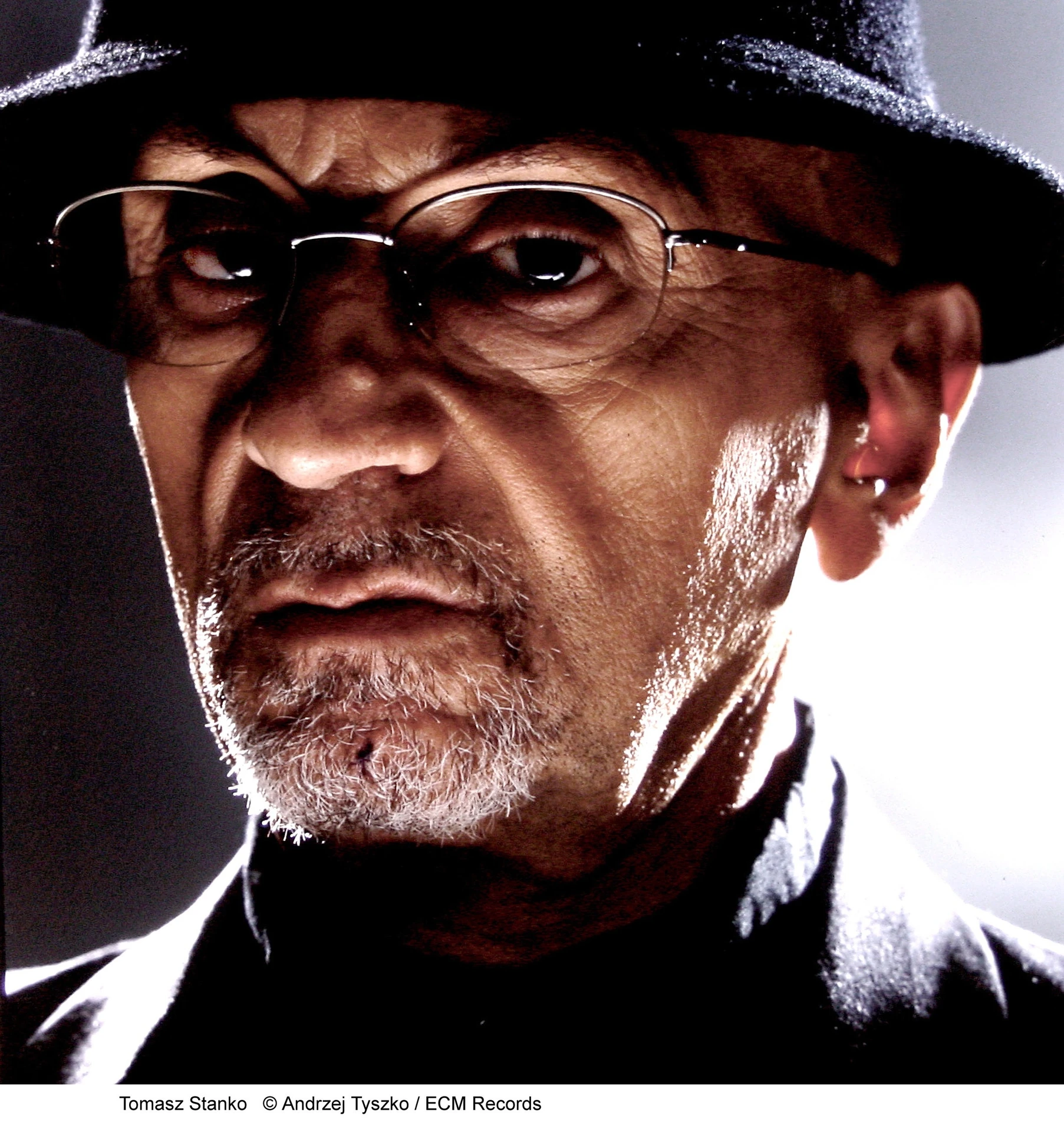 Tomasz Stańko wearing a hat and glasses in a dark portrait
